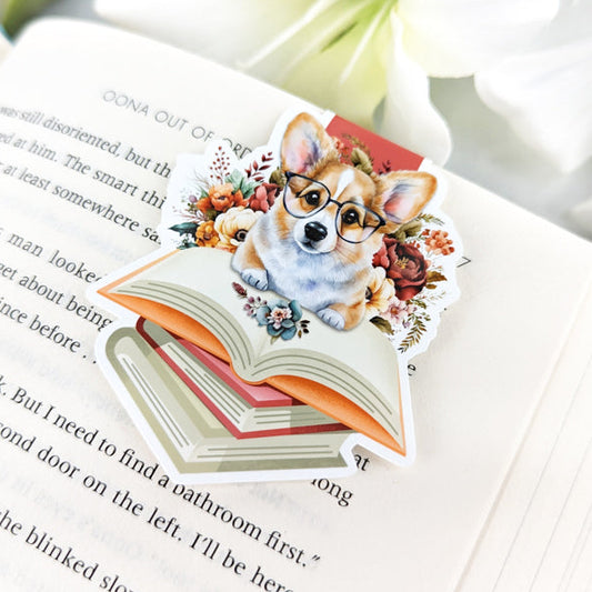 A close-up of a colorful magnetic bookmark featuring a corgi puppy wearing glasses and peeking out from behind a stack of books.