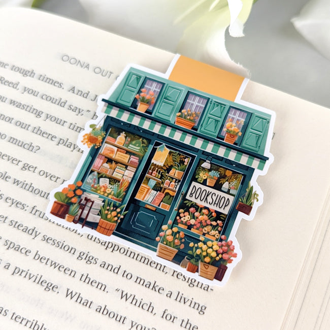 Magnetic bookmark shaped like a floral bookshop with green shutters, orange flowers, and a 'Bookshop' sign, clipped to an open book.