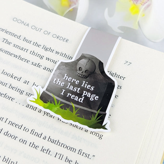 Magnetic bookmark shaped like a tombstone with a skull and the phrase 'Here lies the last page I read,' clipped to an open book.