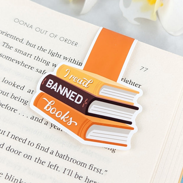 Magnetic bookmark shaped like a stack of orange and brown books with the phrase 'I read banned books,' clipped to an open book.