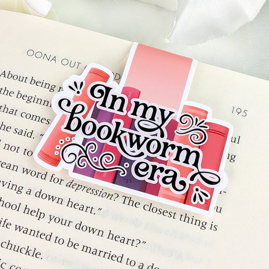 Magnetic bookmark with the phrase “In my bookworm era” in black lettering over a background of pink and purple book spines, clipped onto an open book.