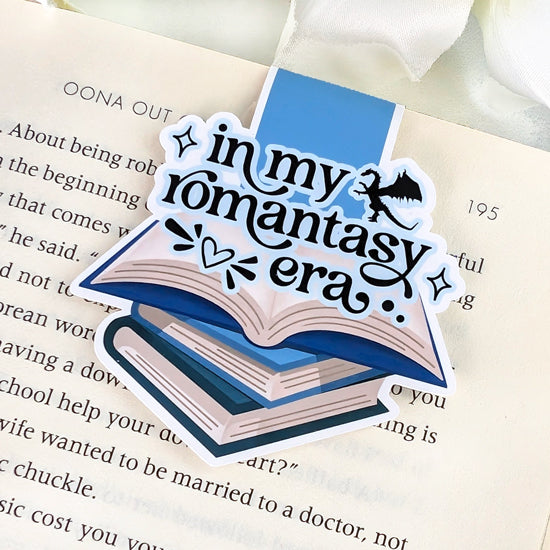 Magnetic bookmark with the phrase “In my romantasy era” in black script above an illustration of open blue-toned books and a small dragon silhouette, clipped onto an open book.