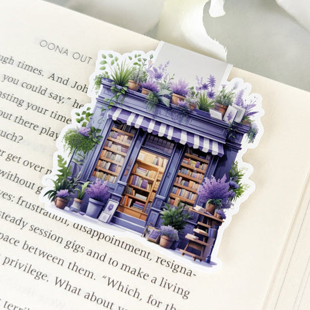 A magnetic bookmark
depicting a charming lavender
bookstore surrounded by lush
greenery, placed on an open
book.