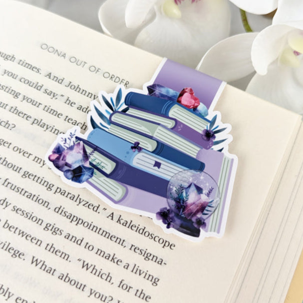 Magnetic bookmark featuring a stack of purple and blue books decorated with crystals, leaves, and sparkles, clipped to an open book.