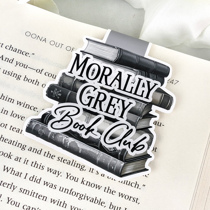 Magnetic bookmark with stacked black and grey books and the phrase "Morally Grey Book Club" in bold text, clipped to an open book.
