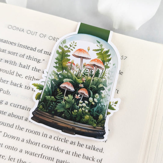 Colorful magnetic bookmark featuring a terrarium with mushrooms and plants inside, tucked into the pages of a book.