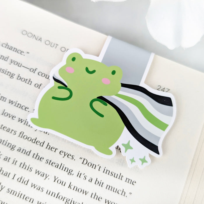 A magnetic bookmark featuring a cute green frog with a smiling face and a flowing Agender Pride Flag cape.