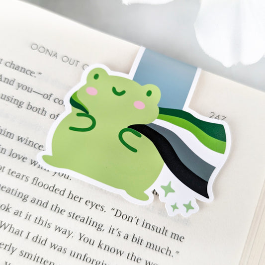 A magnetic bookmark featuring a cute green frog with a smiling face and a flowing Aromantic Pride Flag cape.