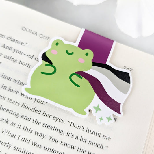 A magnetic bookmark featuring a cute green frog with a smiling face and a flowing Asexual Pride Flag cape.