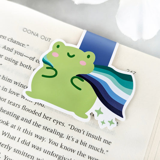 A magnetic bookmark featuring a cute green frog with a smiling face and a flowing Gay Pride Flag cape.