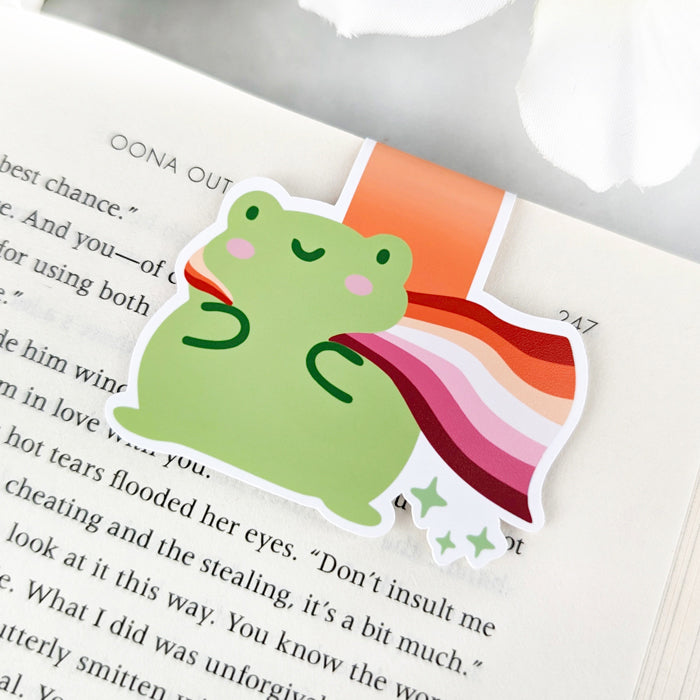 A magnetic bookmark featuring a cute green frog with a smiling face and a flowing Lesbian Pride Flag cape.