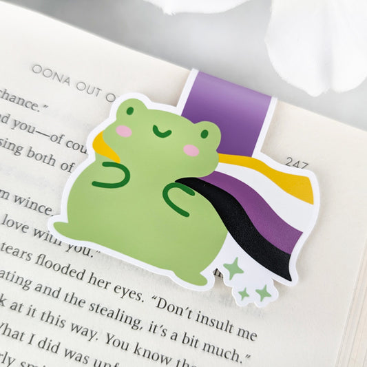 A magnetic bookmark featuring a cute green frog with a smiling face and a flowing Non-Binary Pride Flag cape.