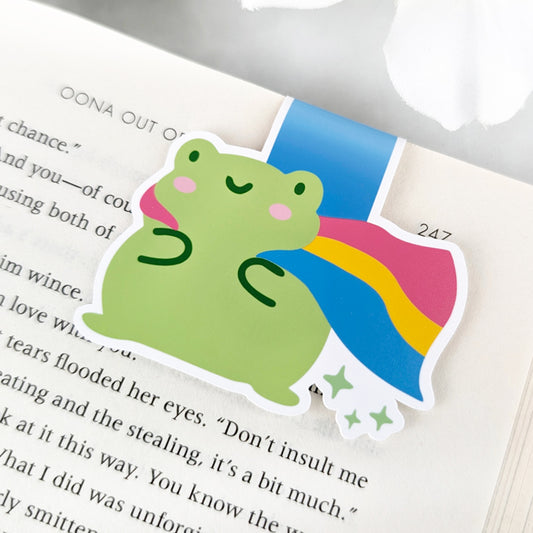 A magnetic bookmark featuring a cute green frog with a smiling face and a flowing Pansexual Pride Flag cape.