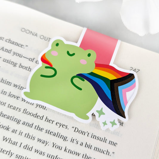 A magnetic bookmark featuring a cute green frog with a smiling face and a flowing Progress Pride Flag cape.