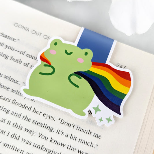 A magnetic bookmark featuring a cute green frog with a smiling face and a flowing rainbow Pride Flag cape.