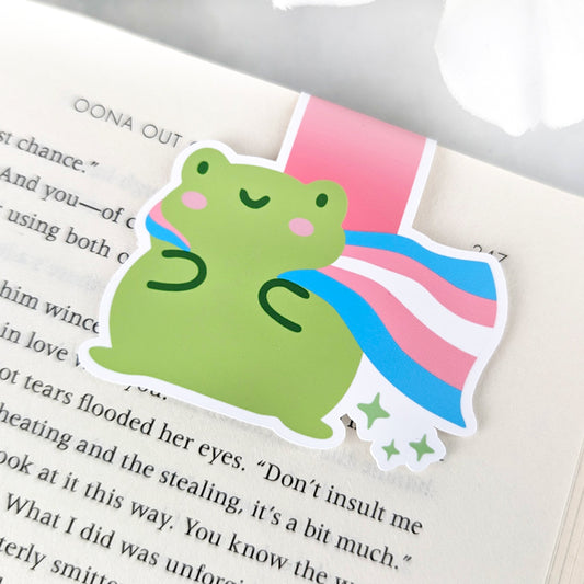 A magnetic bookmark featuring a cute green frog with a smiling face and a flowing Trans Pride Flag cape.