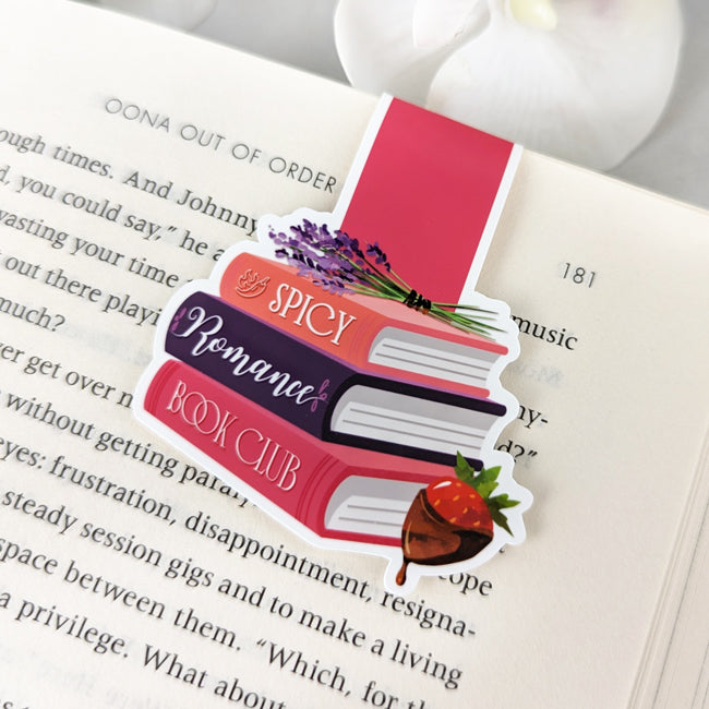 Magnetic bookmark shaped like a stack of romance books labeled 'Spicy Romance Book Club,' with lavender and a chocolate-dipped strawberry, clipped to an open book.