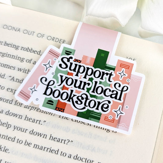 Magnetic bookmark with the phrase “Support your local bookstore” in black script over a background of green and pink books, clipped onto an open book.