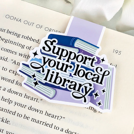 Magnetic bookmark with the phrase “Support your local library” in black lettering, featuring stacked blue and purple books and sparkles, clipped onto an open book.