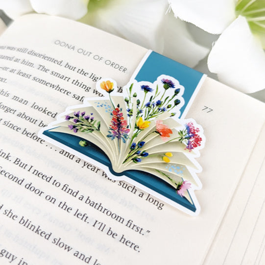 Magnetic bookmark shaped like an open book with colorful wildflowers bursting from the pages, clipped to an open book.