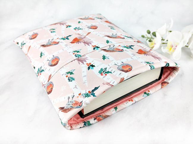 Light pink fabric book sleeve with a birch tree forest pattern featuring robins, holly leaves, and snowflakes, shown holding a hardcover book on a white marble background.