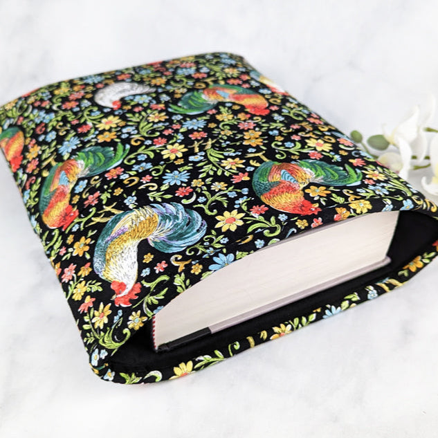 large padded book sleeve with pocket. fabric showcases colorful roosters, hens and folk style flowers on a black background.