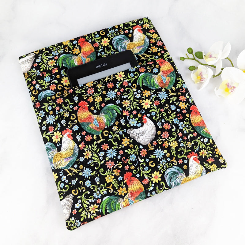 large padded book sleeve with pocket. fabric showcases colorful roosters, hens and folk style flowers on a black background.