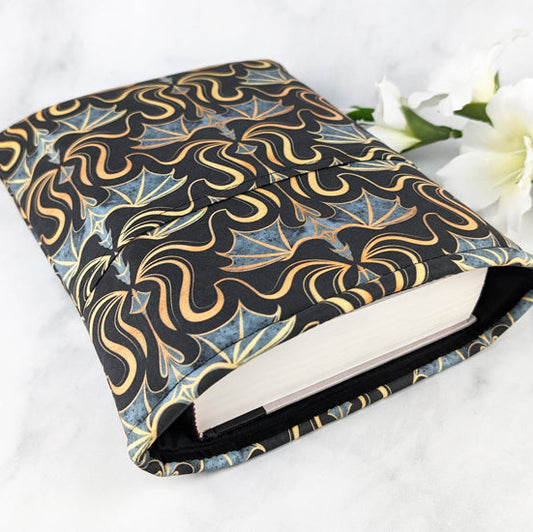 Fabric book sleeve with a bold pattern of gold and gray dragons on a black background, displayed with white flowers on a white surface.
