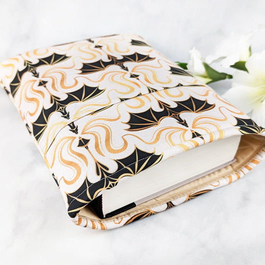 padded large book sleeve with pocket, fabric showcases simple illustrations of flying dragons in a damask style in shades of black, gold and white.