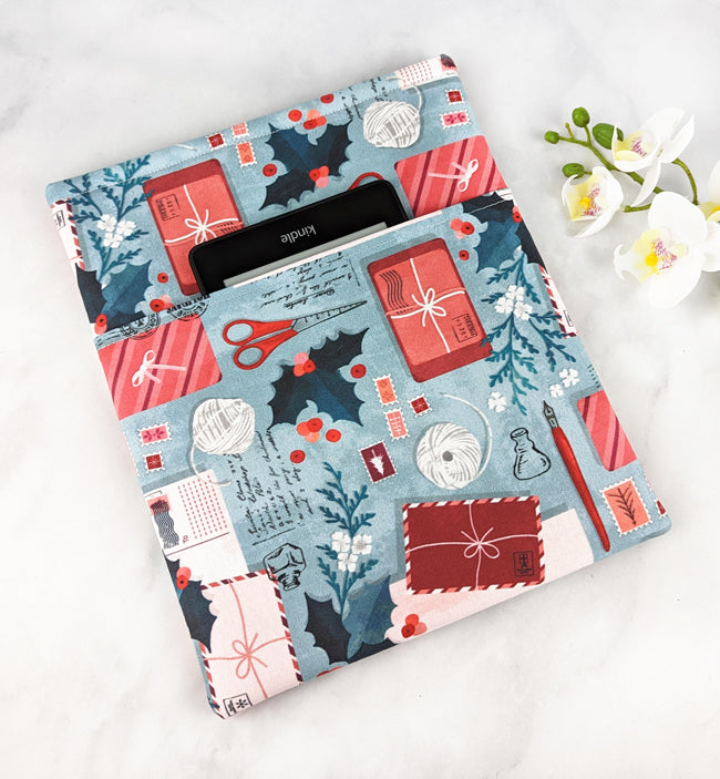 Padded fabric book sleeve with a festive print featuring holiday cards, scissors, holly, and yarn balls on a light blue background, shown holding a hardcover book.