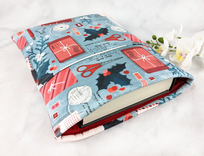 Padded fabric book sleeve with a festive print featuring holiday cards, scissors, holly, and yarn balls on a light blue background, shown holding a hardcover book.