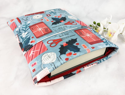 Padded fabric book sleeve with a festive print featuring holiday cards, scissors, holly, and yarn balls on a light blue background, shown holding a hardcover book.