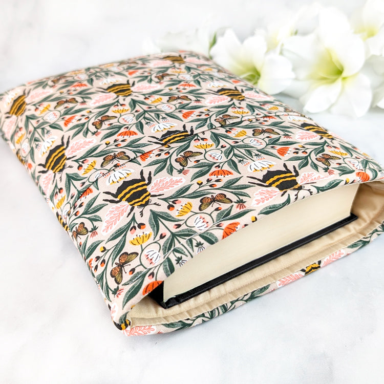 Beige book sleeve with a lively bee and butterfly pattern surrounded by pink, white, and yellow wildflowers on leafy green vines.