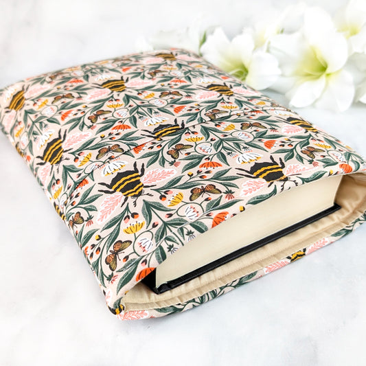 Beige book sleeve with a lively bee and butterfly pattern surrounded by pink, white, and yellow wildflowers on leafy green vines.