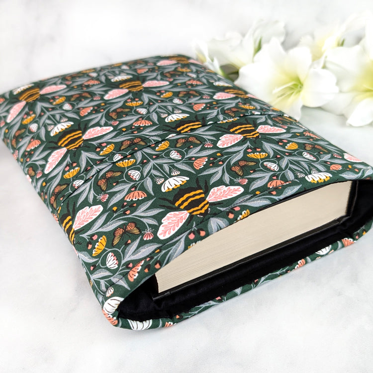Dark green book sleeve with a lively bee and butterfly pattern surrounded by pink, white, and yellow wildflowers on leafy vines.