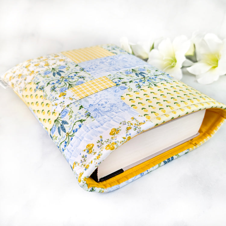Yellow and blue patchwork fabric book sleeve shown from the side with a hardcover book partially inserted and yellow lining visible.