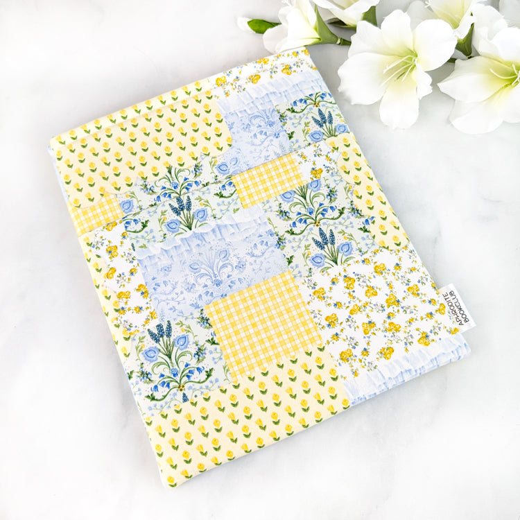 Yellow, blue, and green patchwork fabric book sleeve with small floral patterns, photographed flat on a white marble background with white flowers nearby.
