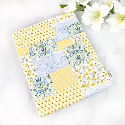 Yellow, blue, and green patchwork fabric book sleeve with small floral patterns, photographed flat on a white marble background with white flowers nearby.