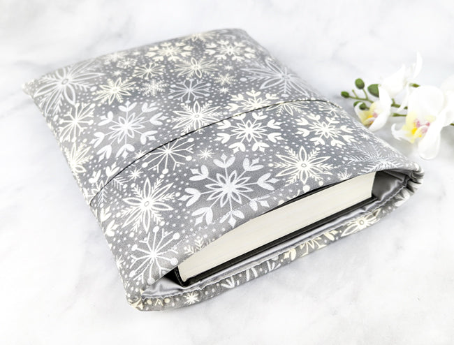 Soft gray fabric book sleeve with a white snowflake pattern, shown holding a hardcover book on a light marble surface with white flowers in the background.