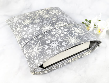 Soft gray fabric book sleeve with a white snowflake pattern, shown holding a hardcover book on a light marble surface with white flowers in the background.
