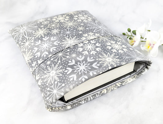 Soft gray fabric book sleeve with a white snowflake pattern, shown holding a hardcover book on a light marble surface with white flowers in the background.