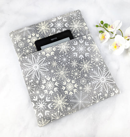 Soft gray fabric book sleeve with a white snowflake pattern, shown holding a hardcover book on a light marble surface with white flowers in the background.