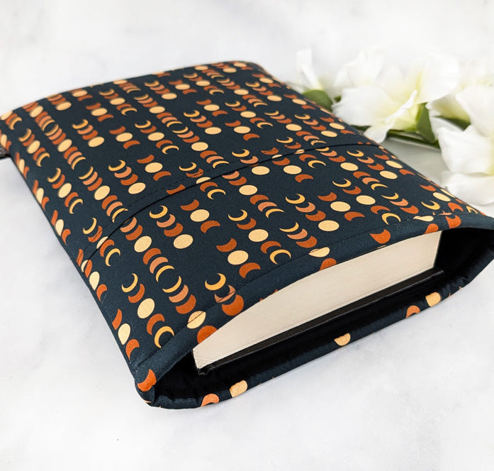 large padded book sleeve with pocket. fabric showcases moon phases in beige and orange shades on a dark navy background.