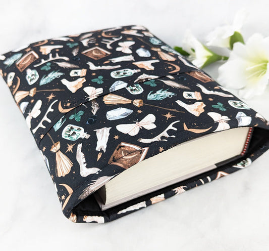 large padded book sleeve with pocket, fabric features various witchy items like crystals, brooms, frogs, moons, spell books etc, on a black background.