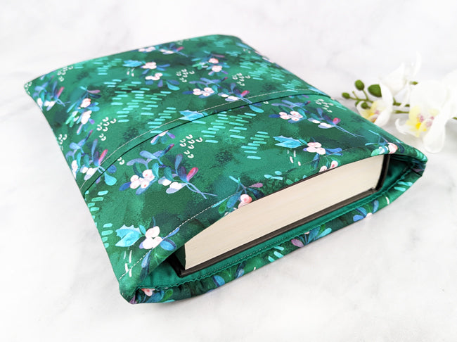 Green fabric book sleeve featuring a mistletoe-inspired floral pattern with soft pink and blue berries, holding a hardcover book on a white marble background.