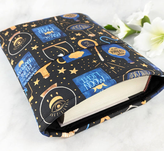 large padded book sleeve with pocket,  fabric pattern features various witchy items like potions, spell books, crystal balls and more. Colors are blue, yellow gold and white on a black background.