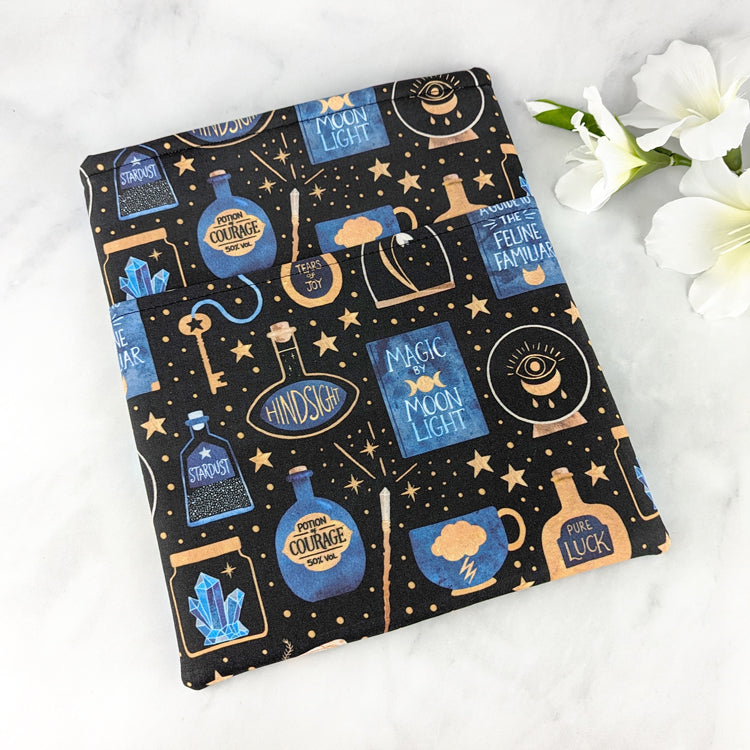 large padded book sleeve with pocket,  fabric pattern features various witchy items like potions, spell books, crystal balls and more. Colors are blue, yellow gold and white on a black background.