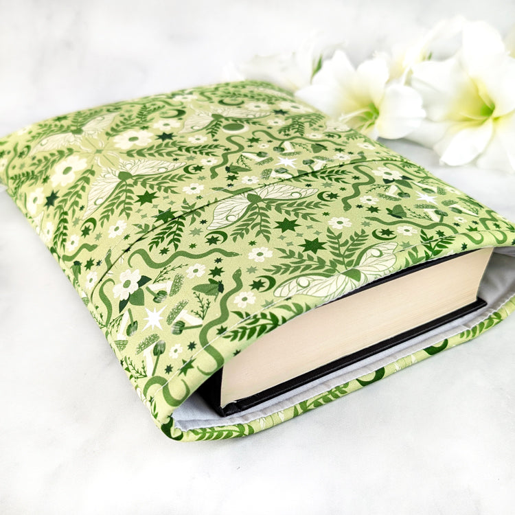Light green book sleeve featuring a celestial moth and floral design with moons, stars, and leafy vines in soft forest tones.