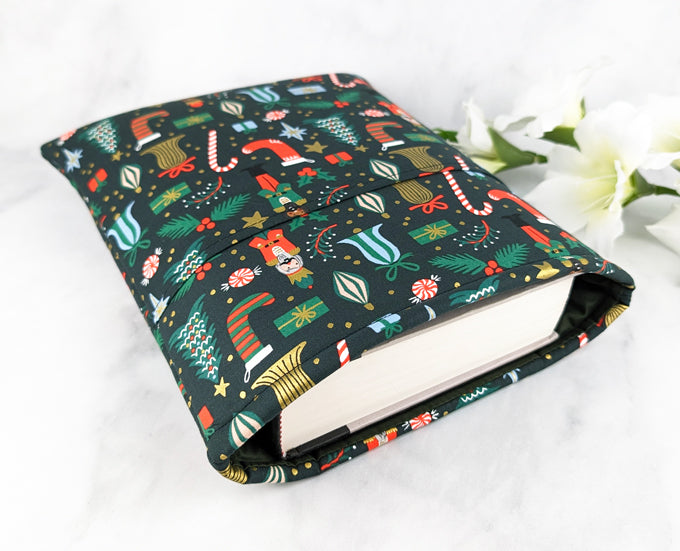 Dark green fabric book sleeve with a festive Nutcracker-themed print featuring candy canes, stockings, ornaments, and gifts, shown holding a hardcover book on a white marble surface with white flowers in the background.