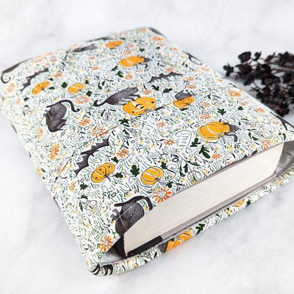 Fabric book sleeve with a cream background and a Halloween-themed pattern of black cats, bats, pumpkins, mushrooms, and orange flowers scattered among green vines.
